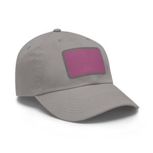 Könnte beinhalten: Graue Baseballkappe mit einem rosa und grauen Aufnäher. Der Aufnäher ist rechteckig und weist diagonale rosa Streifen auf. Die Kappe hat einen gebogenen Schirm und einen Knopf oben.