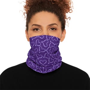 May include: A purple neck gaiter featuring a repeating pattern of white hearts. The face covering is worn around the neck and covers the lower face. The background is white.