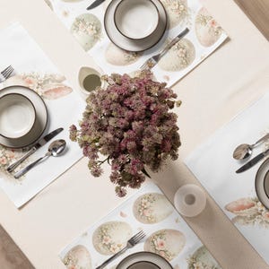 Könnte beinhalten: Draufsicht auf eine Tischdekoration mit Tischsets zum Thema Ostern, Tellern, Schalen und Besteck. Die Tischsets zeigen Eierdesigns mit floralen Akzenten. In der Mitte steht eine Vase mit lila Blumen.