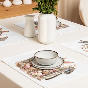 Könnte beinhalten: Eine Tischdekoration mit weißen Tischsets mit einem Ostermotiv. Die Tischsets zeigen ein Nest mit Eiern, Blumen und Hasenohren. Eine weiße Vase mit grünen Stielen, ein Sahnekännchen und eine Schüssel mit einem Teller sind zu sehen.