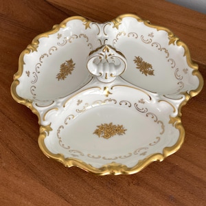 Peut inclure: Un plat de service en porcelaine blanche à trois compartiments et une poignée. Le plat a une bordure dorée et des motifs floraux. Les bords sont festonnés, et le plat est sur une surface en bois.