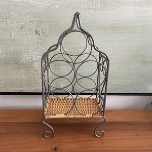 Peut inclure: Un casier à vin en métal gris avec une base en rotin tressé. Le casier a un sommet en forme d'arche et peut contenir plusieurs bouteilles. Le casier est conçu pour être posé sur une table ou un comptoir.