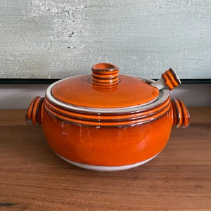 May include: A round ceramic lidded pot with a vibrant orange glaze and dark brown horizontal ring accents. The pot features two side handles and a matching lid with a central knob, plus a small spoon rest opening with a brown-tipped handle.