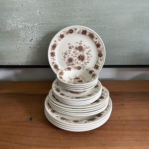 Peut inclure: Une pile de vaisselle en céramique blanche ornée d'un motif floral marron. L'ensemble comprend plusieurs assiettes plates, des assiettes à salade plus petites et un petit bol au sommet. Chaque pièce présente un motif botanique détaillé.