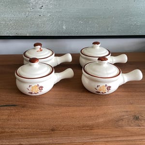 Puede incluir: Cuatro cuencos de sopa de cerámica blanca con tapas y asas. Cada cuenco tiene un borde marrón y un pomo marrón en la tapa. Un diseño floral está en la parte delantera de cada cuenco. Los cuencos están sobre una superficie de madera.