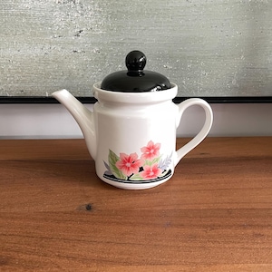 Peut inclure: Théière en céramique blanche avec un couvercle et une poignée noirs. La théière présente un motif floral avec des fleurs roses et des feuilles vertes et bleues. La théière est sur une surface en bois.