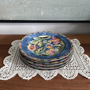May include: A stack of five round ceramic plates featuring a central floral pattern in shades of yellow, pink, green, and blue. The rim is decorated with a multi-coloured chequered border, and the outer edge is a textured, mottled blue finish.