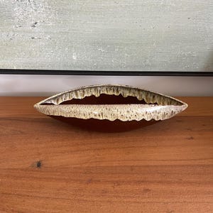 Op de afbeelding: Een langwerpige keramische kom in mid-century modern stijl. De kom heeft een dieprood-bruin interieur en een getextureerde, gebroken witte en bruine rand. De kom staat op een houten oppervlak.