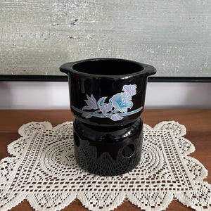 Peut inclure: Un caquelon à fondue en céramique noire avec deux petites anses et une base noire assortie dotée d'ouvertures circulaires. Le pot est orné d'un motif floral dans des tons bleu clair, lavande et blanc, avec des pétales et des feuilles.