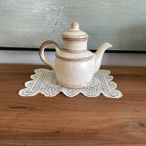 Może przedstawiać: Nakrapiany ceramiczny imbryk z pokrywką i zakrzywionym uchwytem. Imbryk jest w kolorze złamanej bieli z brązowymi akcentami i stoi na białej koronkowej serwetce. Dekoracyjny przedmiot do kuchni lub jadalni.