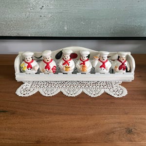 Peut inclure: Un ensemble de six figurines de chefs en céramique disposées dans un support en bois blanc. Chaque chef porte un uniforme blanc avec un foulard rouge et une toque. Le support présente des illustrations peintes d'aubergine, tomate et poivron.