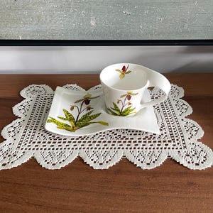 Może przedstawiać: Biały ceramiczny zestaw składający się z filiżanki i prostokątnego spodka, ozdobiony botaniczną ilustracją storczyka obuwika z zielonymi liśćmi i kasztanowymi kwiatami. Filiżanka posiada szerokie, zakrzywione ucho, a spodek falisty brzeg.