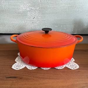 Peut inclure: Une cocotte ovale en fonte émaillée arborant un dégradé de couleur orange vif. Le récipient est doté de deux poignées latérales, d'un couvercle assorti avec un motif de rebord circulaire et d'un petit bouton noir centré sur le dessus. La finition extérieure passe d'un rouge-orange profond à la base à un orange plus clair et lumineux vers le bord supérieur.