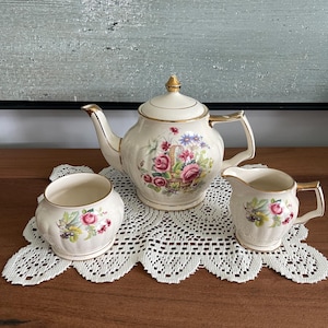 Könnte beinhalten: Ein cremefarbenes Porzellan-Teeservice bestehend aus einer Teekanne mit Deckel, einer Zuckerdose und einem Milchkännchen. Jedes Teil zeigt ein Blumenmuster aus rosa Rosen und blauen Blüten mit Goldrand an den Rändern, Griffen und dem Deckelknauf.