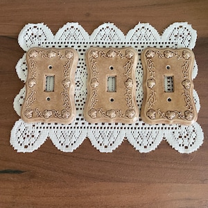 Peut inclure: Trois plaques d'interrupteur rectangulaires en céramique beige, chacune présentant un motif en relief de vignes fleuries le long de la bordure. Les plaques ont une découpe centrale rectangulaire pour un interrupteur et deux petits trous de vis.