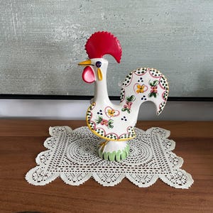 Puede incluir: Una figura de gallo de cerámica con cuerpo blanco y patrones florales pintados a mano en rosa, amarillo y verde. El gallo tiene una cresta roja prominente, un pico amarillo y una carúncula roja, sobre una base decorada con motivos de hojas verdes.