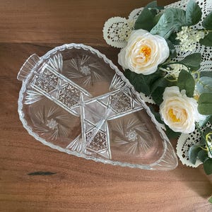 May include: A clear glass serving dish shaped like a leaf, featuring intricate cut patterns including starburst motifs and geometric cross-hatching. The dish has a serrated edge and a textured surface, designed with a central divider creating two sections.