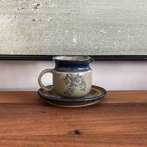 Peut inclure: Un ensemble de pot à crème et soucoupe en céramique. Le pot à crème est beige avec un bord et une anse bleus, et un motif floral bleu. La soucoupe est beige avec un bord bleu. L'ensemble est sur une surface en bois.