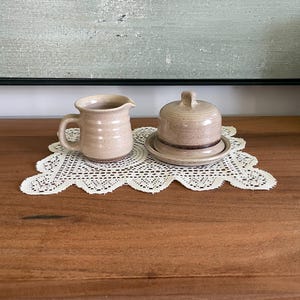Peut inclure: Un petit pot à crème en céramique beige avec une anse et un beurrier assorti avec couvercle. Les deux sont placés sur un napperon en dentelle blanche. Les articles sont sur une surface en bois, avec une peinture en arrière-plan.