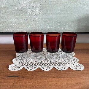 Könnte beinhalten: Vier rubinrote Weingläser mit klaren Stielen und Böden sind auf einer weißen Spitzendecke angeordnet. Die Gläser haben eine kurze, zylindrische Form und eine dunkelrote Farbe, die nach oben hin verblasst. Die Gläser stehen auf einer Holzoberfläche.