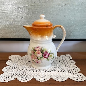 Peut inclure: Une cafetière en porcelaine avec une base blanche et un dégradé orange vif sur la partie supérieure et le couvercle. Le corps est orné d'un arrangement floral détaillé de roses roses, jaunes et blanches, complété par une anse et un bec verseur.