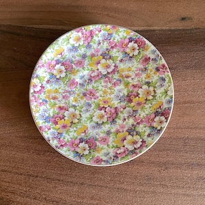 May include: A round, floral-patterned plate with a gold rim. The plate features a dense pattern of pink, yellow, white, and blue flowers on a cream background. The plate is likely made of ceramic or porcelain.