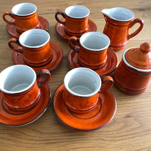 Peut inclure: Un ensemble de tasses à expresso en céramique orange avec soucoupes assorties, une crémière et un sucrier avec couvercle. Les tasses ont des intérieurs blancs et des anses marron foncé. L'ensemble est disposé sur une surface en bois.