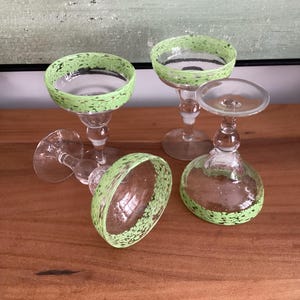 May include: Four clear margarita glasses with green speckled rims. The glasses are on a wooden surface, with one glass lying on its side. The stem of each glass has a round bulb shape.