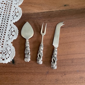 May include: Set of three silver cheese serving utensils. Includes a pie server, a two-pronged fork, and a cheese knife. The handles are decorated with a grape vine design. The utensils are displayed on a wooden surface.