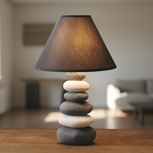 May include: A table lamp featuring a base made of stacked stones in grey and white hues. It has a dark brown, conical lampshade. The lamp sits on a wooden table, with a blurred room in the background.
