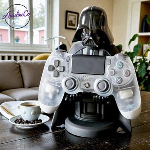 Może przedstawiać: Czarny stojak na kontroler do gier w stylu Darth Vadera trzymający przezroczysty kontroler do gier. Kontroler ma białe akcenty i kolorowe przyciski. Filiżanka kawy stoi na spodku po lewej stronie stojaka.