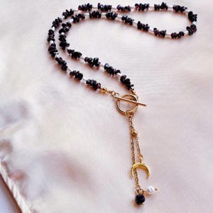 Op de afbeelding: Een delicate ketting met donkerrode edelsteen chips en kleine witte kralen. De ketting heeft een goudkleurige toggle sluiting en een bungelende bedel met een halve maan, een parel en een donkerrode kraal.