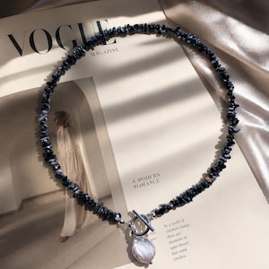 May include: A necklace composed of small, irregular black and white stone chips strung together, featuring a silver-toned toggle clasp at the front. A single, large, iridescent white baroque pearl pendant hangs from the center of the toggle closure.