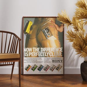 May include: Framed vintage Sony advertisement featuring a cassette tape and a yellow portable player. The poster has the text "NOW THE DIFFERENCE IS PERFECTLY CLEAR." Several cassette tape designs are shown at the bottom.