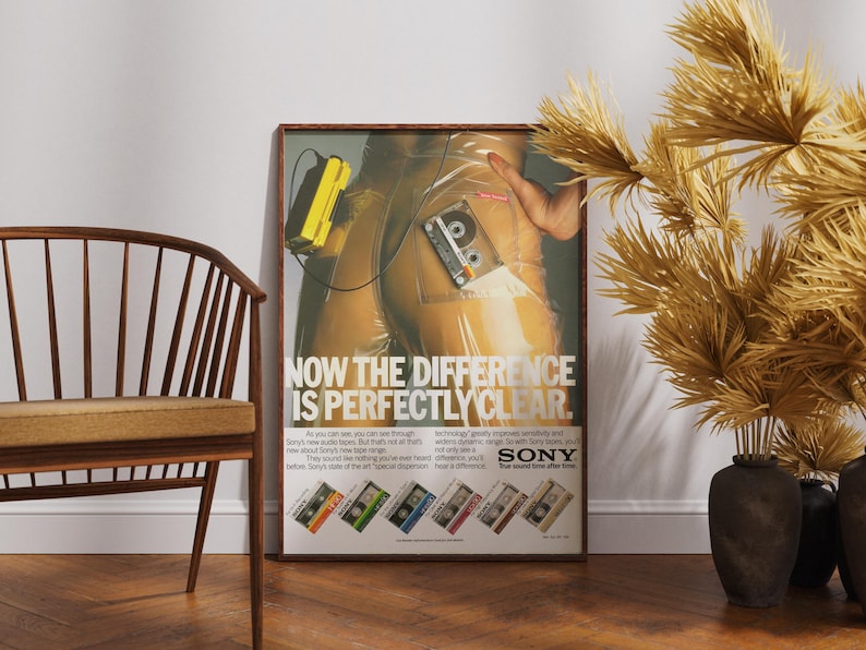 May include: Framed vintage Sony advertisement featuring a cassette tape and a yellow portable player. The poster has the text "NOW THE DIFFERENCE IS PERFECTLY CLEAR." Several cassette tape designs are shown at the bottom.