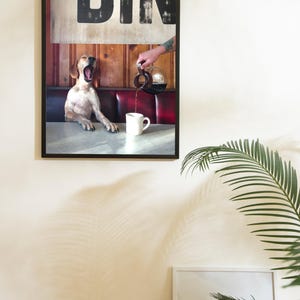 May include: Framed print featuring a dog yawning in a diner booth as coffee is poured into a mug. The diner sign is visible above the booth. The image has a vintage aesthetic with a red and wood-paneled booth.