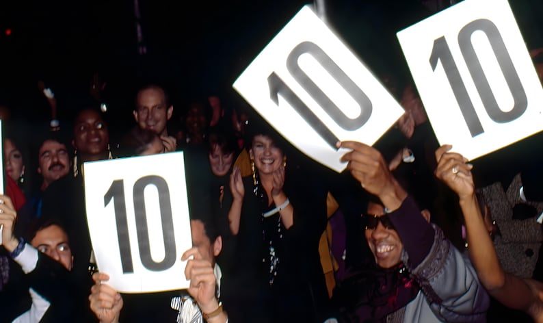 May include: A crowd of people holding up signs with the number 10 on them. The people are smiling and appear to be enjoying themselves. The image is taken in a dimly lit room.