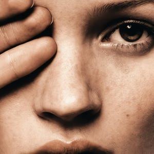 May include: Close-up of a person's face, with a hand partially covering the eye. The skin tone is light brown, and the eye is dark brown. The image is well-lit, highlighting the textures of the skin and the details of the face.