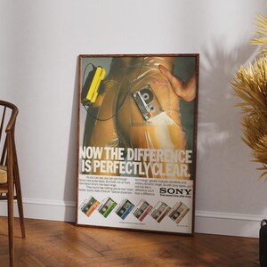 May include: A framed vintage advertisement for Sony audio cassettes. The poster features a retro design with a person and a cassette tape. The text reads "NOW THE DIFFERENCE IS PERFECTLY CLEAR." The poster is leaning against a white wall.