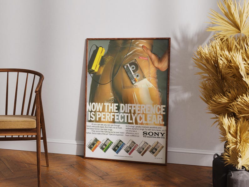May include: A framed vintage advertisement for Sony audio cassettes. The poster features a retro design with a person and a cassette tape. The text reads "NOW THE DIFFERENCE IS PERFECTLY CLEAR." The poster is leaning against a white wall.
