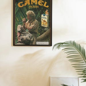 May include: Framed vintage advertisement for Camel Filters cigarettes. The poster features a man smoking a cigarette, a pack of cigarettes, and the Camel Filters logo. The background is a tropical scene with palm leaves.