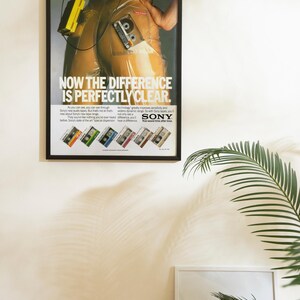 May include: Framed vintage advertisement for Sony audio tapes. The poster features a person in a clear plastic suit with a cassette tape. Text reads "NOW THE DIFFERENCE IS PERFECTLY CLEAR" with the Sony logo and tape examples.