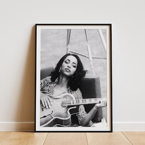 May include: Monochrome framed print featuring a woman with a guitar. She is seated, wearing a patterned top, and has her eyes closed. The guitar is light-coloured with a dark pickguard. The print is leaning against a wall.