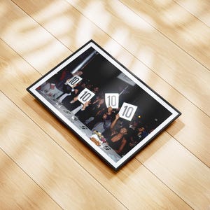 May include: A framed photograph featuring a group of people holding up signs with the number "10" on them. The photograph is in a black frame and is set against a light-colored wooden surface.