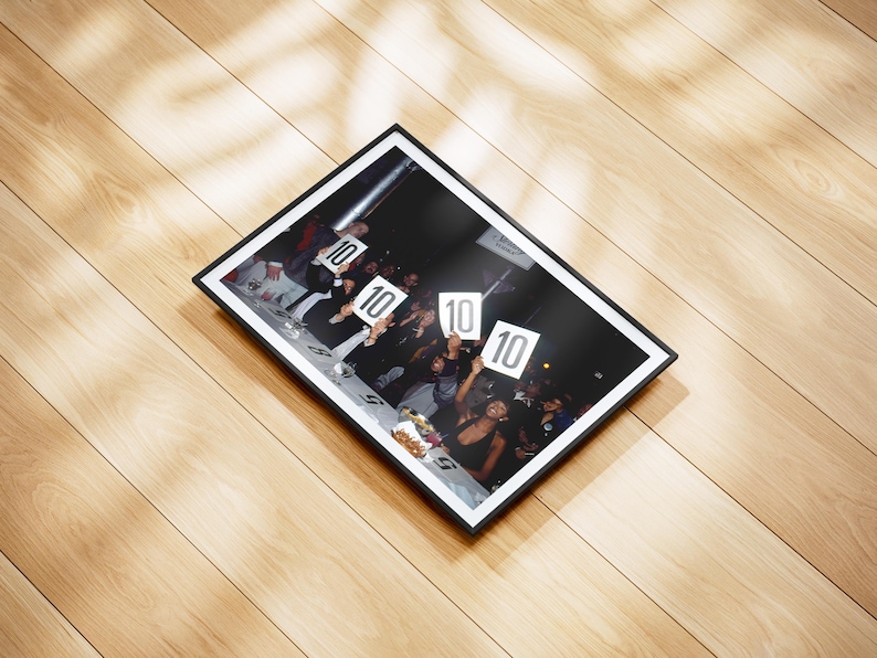 May include: A framed photograph featuring a group of people holding up signs with the number "10" on them. The photograph is in a black frame and is set against a light-colored wooden surface.