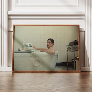 Puede incluir: Impresión enmarcada que representa a una persona en una bañera, fumando un cigarro y leyendo un libro. El baño tiene una estética vintage, con una pared alicatada y una pequeña mesa auxiliar. La impresión está en un marco de madera.