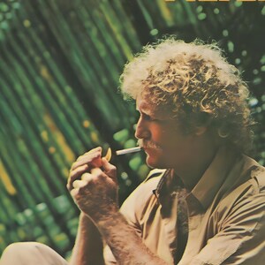 May include: A vintage advertisement poster featuring a person with curly blonde hair and a mustache, wearing a tan button-down shirt. The person is holding a lit cigarette to their lips. The text CAMEL FILTER is visible in yellow.