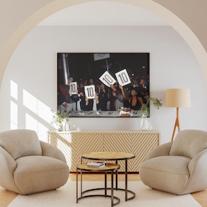 May include: A framed photograph of a group of people holding up signs with the number 10 on them. The photograph is hanging above a wooden cabinet with two beige armchairs and two small round tables in front of it.