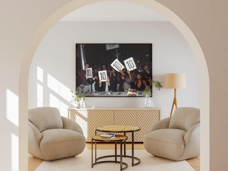 May include: A framed photograph of a group of people holding up signs with the number 10 on them. The photograph is hanging above a wooden cabinet with two beige armchairs and two small round tables in front of it.