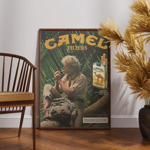 May include: A framed vintage Camel Filters advertisement featuring a man smoking, with the brand's logo and a pack of cigarettes. The poster is set against a white wall, with a wooden chair and decorative plants.